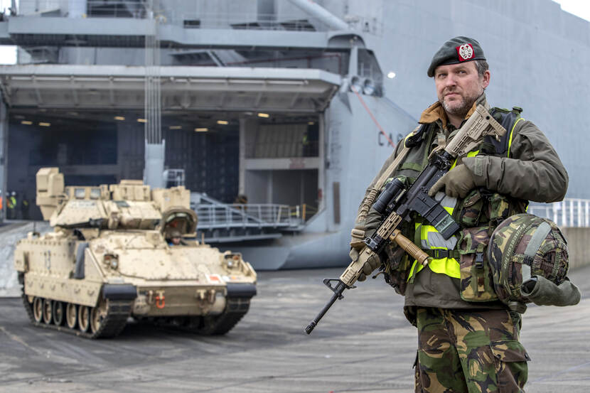 Dutch soldier with weapon stands in front of an American infantry fighting vehicle, with a naval ship in the background.