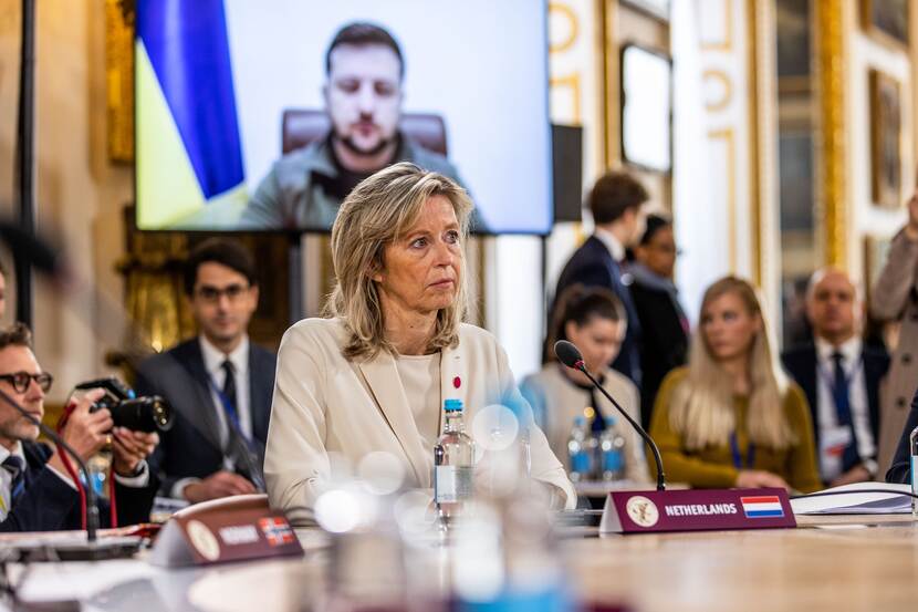 Minister Ollongren aan vergadertafel. Achter haar op een scherm de Oekraïense president.